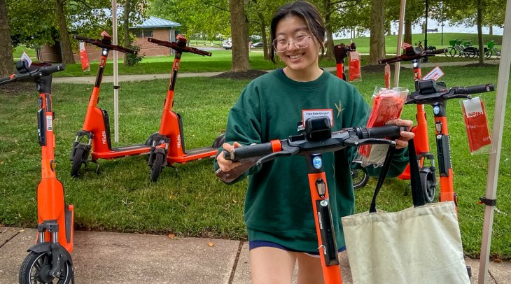 A woman stands under a red SPIN-branded canopy, smiling while holding an orange SPIN electric scooter. She's wearing glasses, a green sweatshirt, shorts, and sandals, and has a beige tote bag on her arm. Behind her, several more scooters are lined up on g